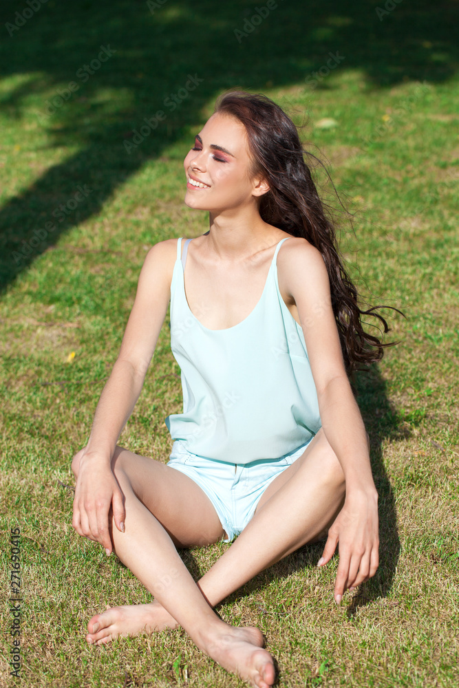 Young brunette woman in a turquoise blouse posing sitting on a green lawn in a summer park Young brunette woman in a turquoise blouse posing sitting on a green lawn in a summer park