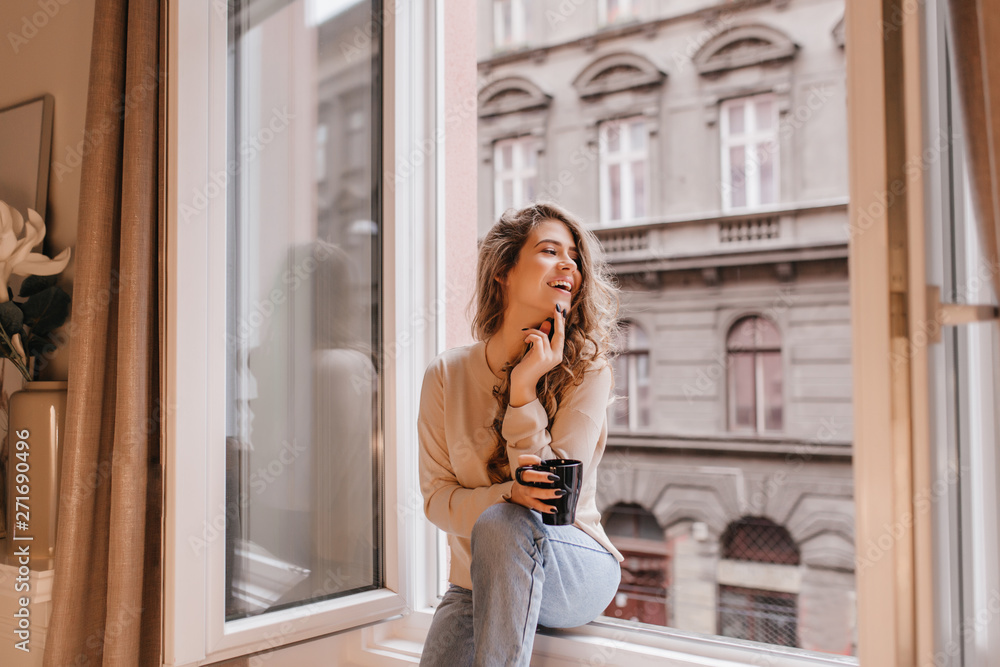 Adorable young woman in trendy jeans enjoying leisure time at home with cup of hot chocolate. Indoor portrait of positive curly girl, sitting on window sill and laughing.