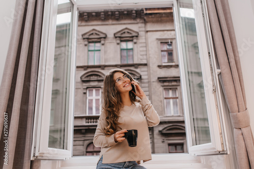 Beautiful glad white lady relaxing in morning with cup of hot beverage. Inspired girl with long hair standing next to opened window and drinking tea with smile.
