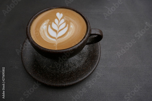 Cappuccino with artful milk foam pattern in a rustic brown cup with saucer. Close-up in front of dark background