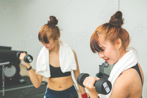 Asian young girl doing exrecises with dumbbell in gym,looking her body through mirror at morning.