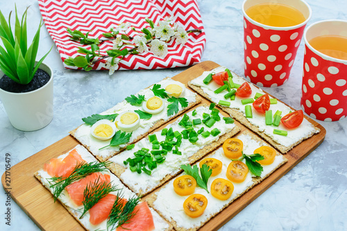 A light summer snack is cooked on a gray table. Small sandwiches with curd cheese, tomatoes and trout. Drinks in bright glasses. Delicious fresh food.