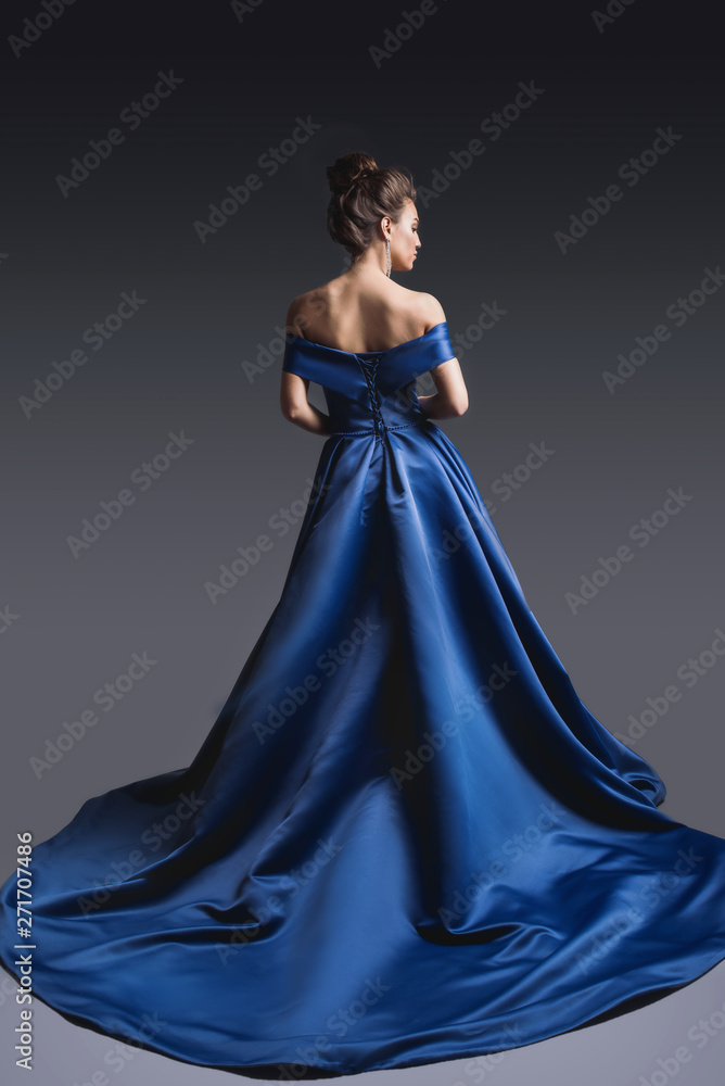 Beautiful woman in elegant blue dress with plume posing on dark background. Back view