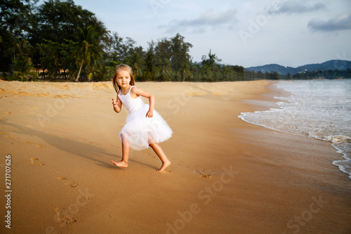 Cute toddler girl with blonde hair in a white tutu dress running on a sandy beach at sunset. Happy childhood memories  Summer vacations   travel concept