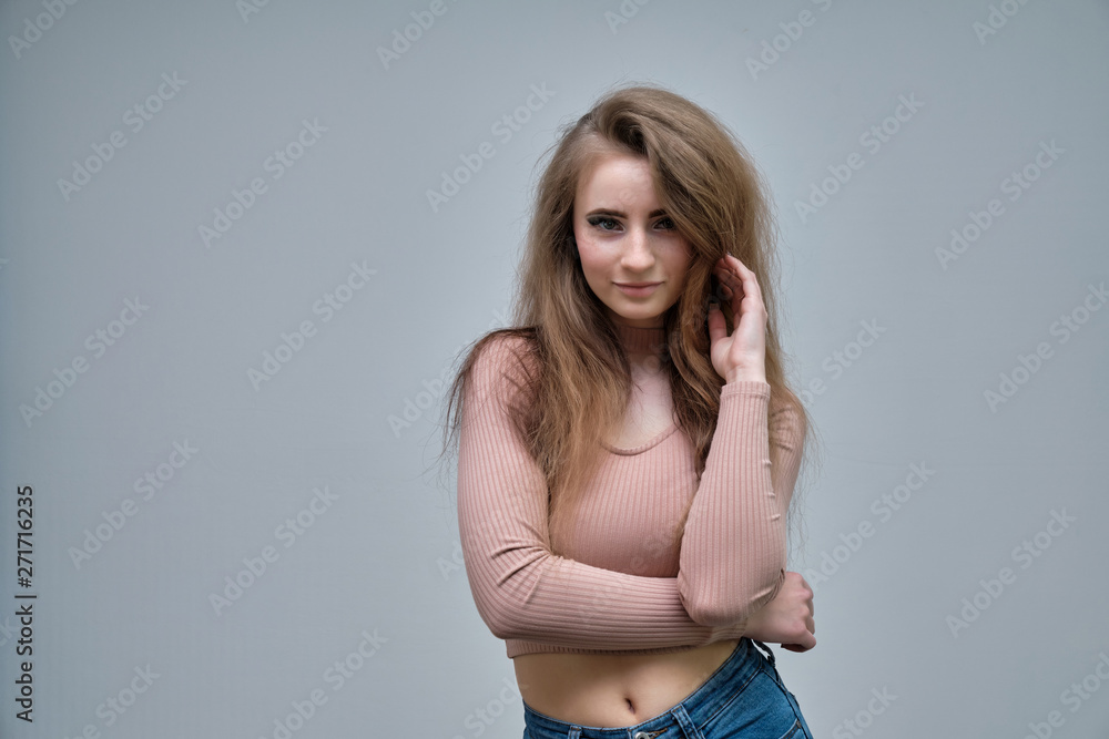 A photo portrait of a beautiful woman girl on a light gray background with long beautiful hair in a pink sweater and blue jeans is standing right in front of the camera and smiling. A photo portrait of a beautiful woman girl on a light gray background with long beautiful hair in a pink sweater and blue jeans is standing right in front of the camera and smiling.