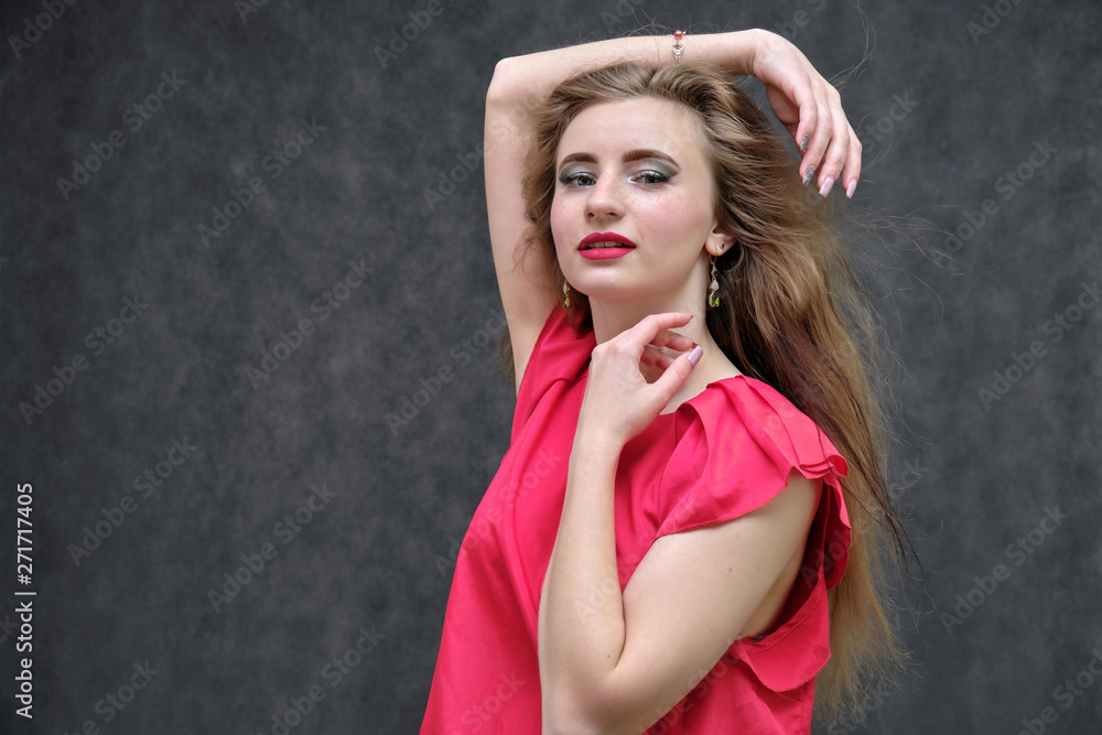Photo portrait of a beautiful girl of the woman on a dark gray background with long beautiful hair in a red sleeveless jacket standing right in front of the camera and smiling. Photo portrait of a beautiful girl of the woman on a dark gray background with long beautiful hair in a red sleeveless jacket standing right in front of the camera and smiling.