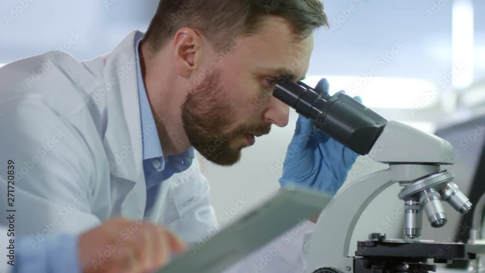 Close up of male and female biochemists in medical gowns standing at desktop in lab. Man working on microscope while his female laboratory assistant writing down results using tablet Close up of male and female biochemists in medical gowns standing at desktop in lab. Man working on microscope while his female laboratory assistant writing down results using tablet