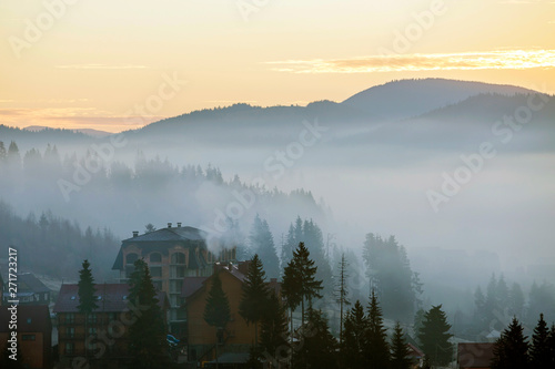 Resort village houses buildings on background of foggy blue mountain hills covered with dense misty spruce forest under bright pink sky at sunrise. Mountain landscape at dawn.