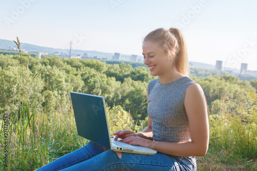 Young girl smiling in nature with laptop on her lap. The girl enjoys looking at the monitor laptop. Close-up portrait.