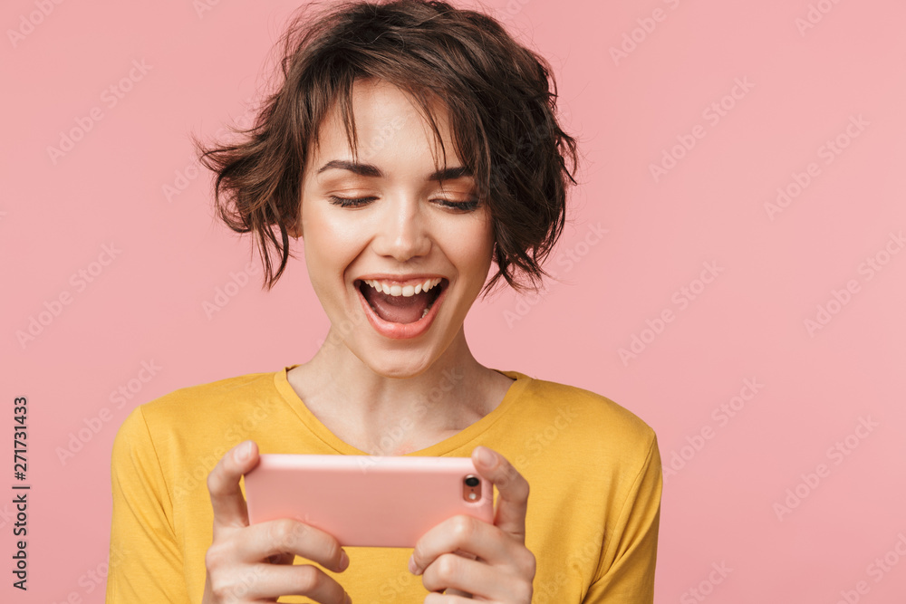 Happy young beautiful woman posing isolated over pink wall background play games by mobile phone. Happy young beautiful woman posing isolated over pink wall background play games by mobile phone.