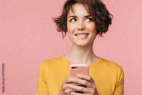Thinking dreaming young beautiful woman posing isolated over pink wall background using mobile phone.