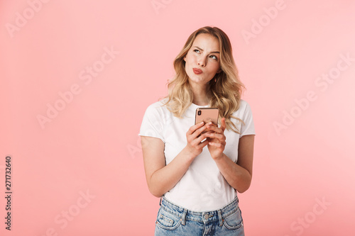 Fotografía Thinking young blonde woman posing isolated over pink wall background using mobile phone