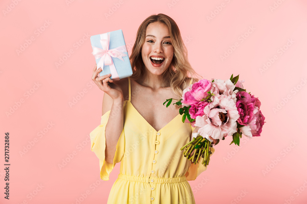 Young blonde woman posing isolated over pink wall background holding flowers holding present gift box. Young blonde woman posing isolated over pink wall background holding flowers holding present gift box.