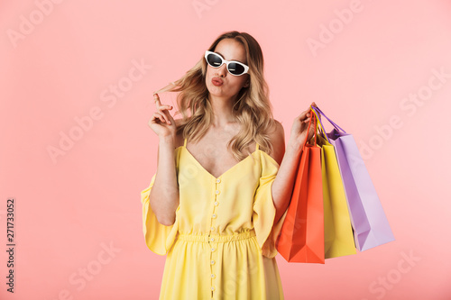 Happy young blonde woman posing isolated over pink wall background holding shopping bags.