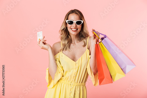 Beautiful happy young blonde woman posing isolated over pink wall background holding shopping bags and credit card.