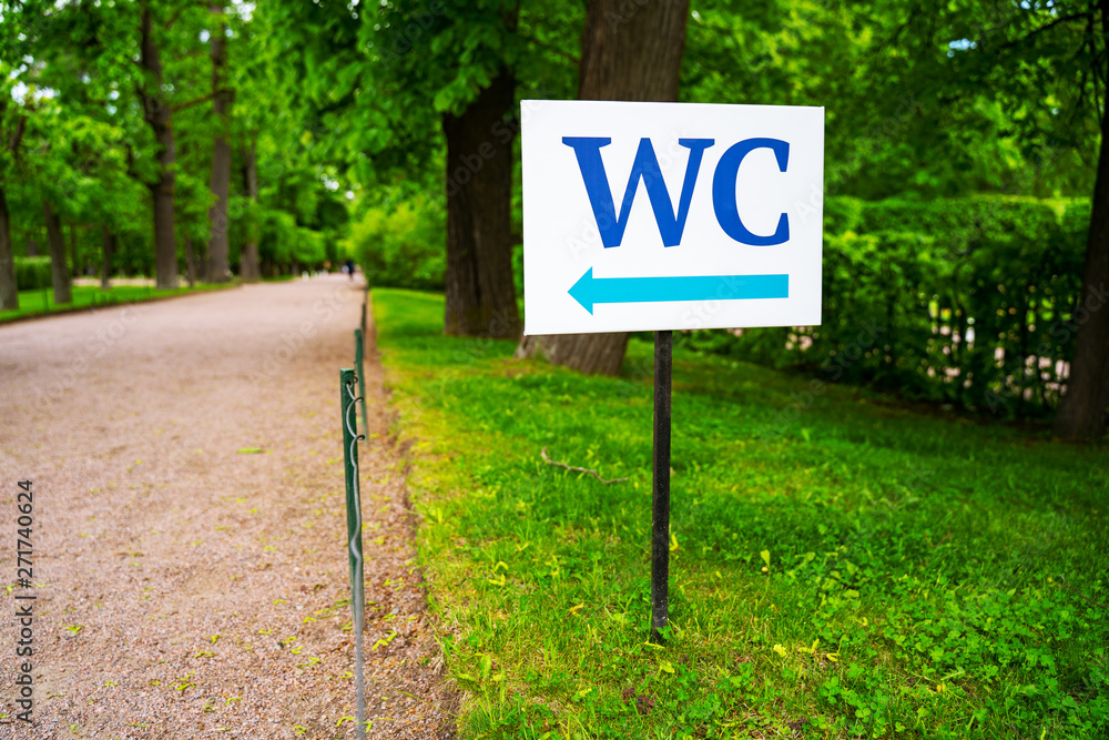 toilet sign against the background of green trees in the park. White WC sign on white metal plate with blue pointing arrow indicating the direction toilet sign against the background of green trees in the park. White WC sign on white metal plate with blue pointing arrow indicating the direction