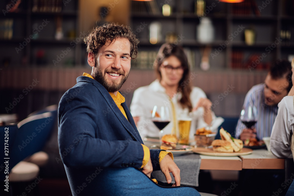 Young smiling man dressed smart casual sitting at restaurant and looking over shoulder. In background his friends having dinner.