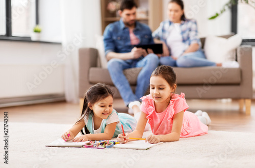 family  leisure and childhood concept - happy sisters lying on floor and drawing in sketchbooks with crayons at home