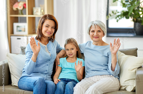 family, generation and female concept - portrait of smiling mother, daughter and grandmother sitting on sofa and waving hands at home