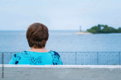 An elderly woman is sitting on a bench. She looks at the sea shore. A beacon is far away on the horizon. She turned her back into the camera. The woman has dark hair, short hair