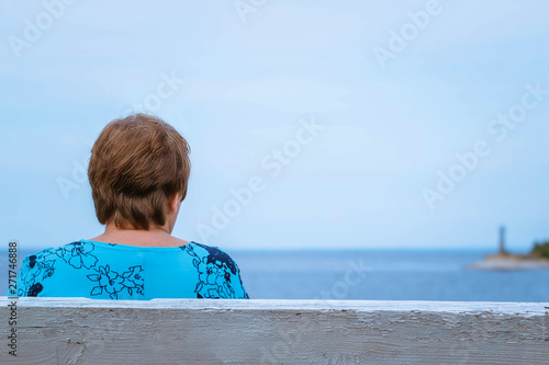 An elderly woman is sitting on a bench. She looks at the sea shore. A beacon is far away on the horizon. She turned her back into the camera. The woman has dark hair, short hair