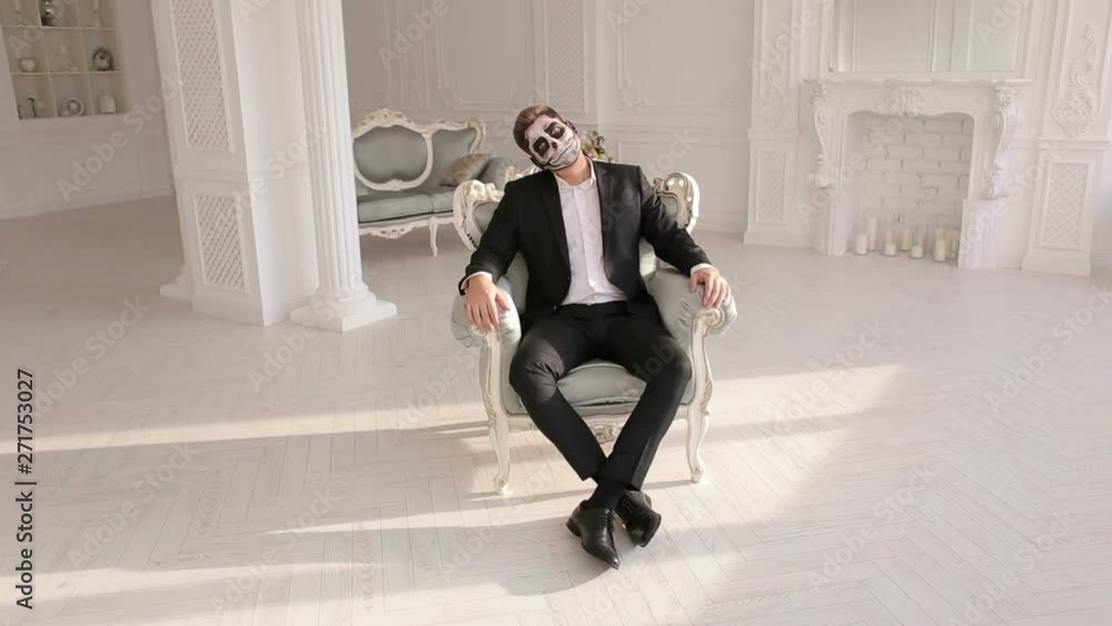 Businessman with make-up skeleton sitting in the old grey chair, and creepy looking at the camera. Halloween or horror theme. Dead face make up portrait of young man in studio. Businessman with make-up skeleton sitting in the old grey chair, and creepy looking at the camera. Halloween or horror theme. Dead face make up portrait of young man in studio.