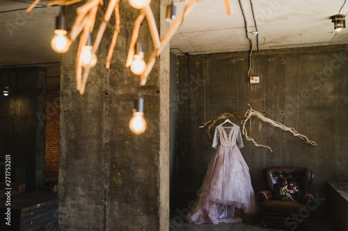 Modern bridal fashion. Lilac wedding dress hanging on a vintage branch inside the concrete loft interior. Bouquet of flowers on a leahter armchair next to it. photo