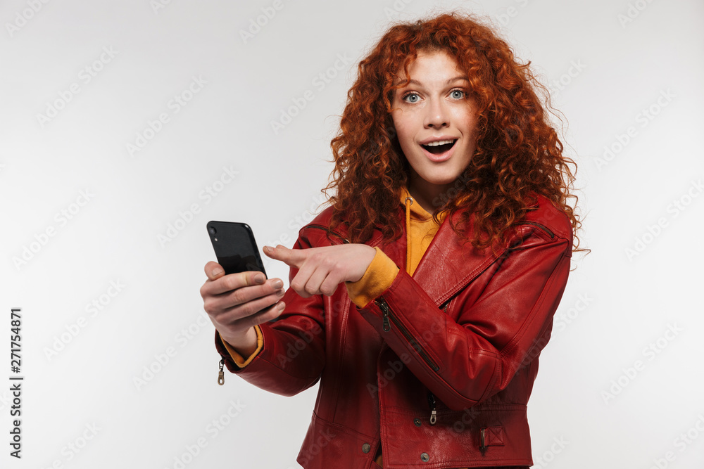 Naklejka premium Image of excited redhead woman 20s wearing leather jacket smiling and holding smartphone