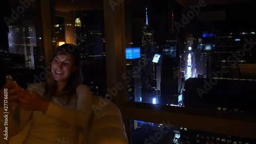 CLOSE UP: Girl points at Times Square while video calling friends at night. Young Caucasian woman skypes with her family from a luxury hotel room with a spectacular view of the New York's cityscape. photo