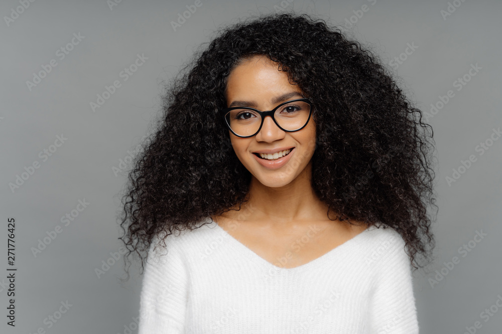 Gorgeous lovely curly woman with Afro hairstyle, feels glad, smiles gently at camera, wears optical glasses and white sweater, isolated on grey background. Happy emotions and feelings concept - obrazy, fototapety, plakaty 