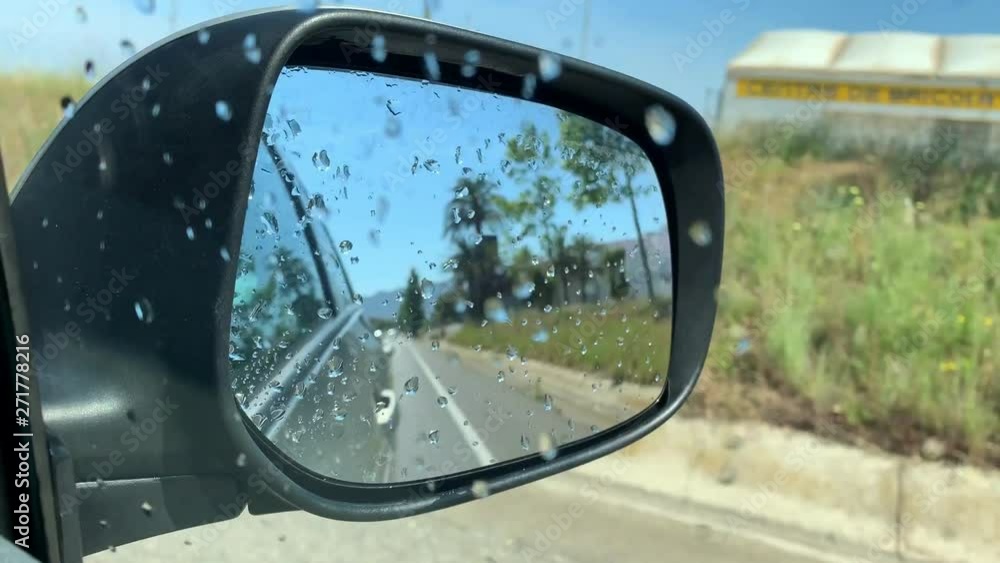 Passenger POV looking through a wet car window in sunny day. Shoot in rear-view mirror of vehicle while driving through the road. Travel in summer day. Water drops on the windows after a car wash. 