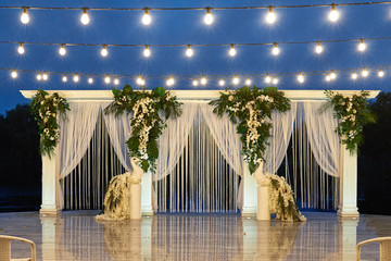 Night wedding ceremony with arch, orchid flowers, palm leaves, chairs and bulb lights in forest outdoors, copy space. Wedding floral decorations