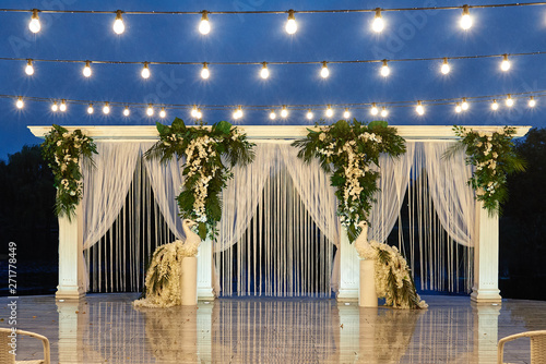 Night wedding ceremony with arch, orchid flowers, palm leaves, chairs and bulb lights in forest outdoors, copy space. Wedding floral decorations