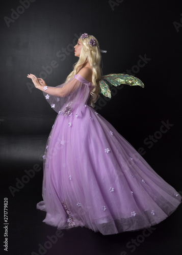  full length portrait of a blonde girl wearing a fantasy fairy inspired costume, long purple ball gown with fairy wings, standing pose on a dark studio background.
