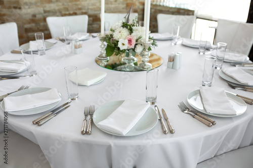 Wedding table setting decorated with fresh flowers. White plates, silverware, white tablecloth and white room. Wedding floristry. Banquet table for guests. Bouquet with roses, hydrangea and eustoma © Kate