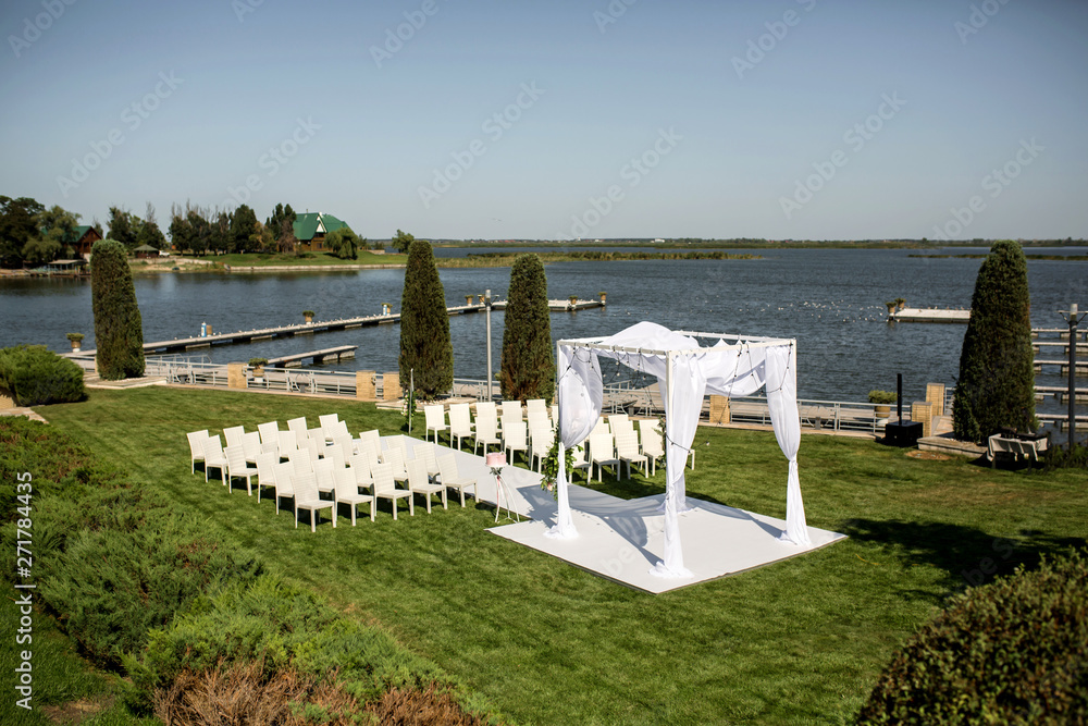 Beautiful outgoing wedding set up. Jewish Hupa on romantic wedding ceremony , wedding outdoor on the lawn water view. Wedding decor. White wooden chairs on a green lawn. White armchairs for guests Beautiful outgoing wedding set up. Jewish Hupa on romantic wedding ceremony , wedding outdoor on the lawn water view. Wedding decor. White wooden chairs on a green lawn. White armchairs for guests