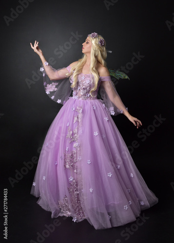  full length portrait of a blonde girl wearing a fantasy fairy inspired costume, long purple ball gown with fairy wings, standing pose on a dark studio background.