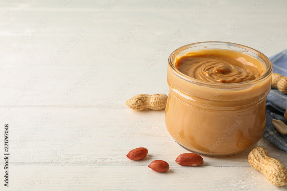 Glass jar with peanut butter, peanut and kitchen towel on white wooden background, closeup and space for text Glass jar with peanut butter, peanut and kitchen towel on white wooden background, closeup and space for text