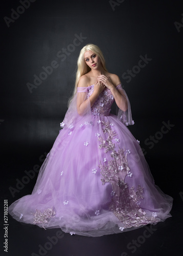 full length portrait of a blonde girl wearing a fantasy fairy inspired costume,  long purple ball gown with fairy wings,   sitting pose  on a dark studio background. © faestock