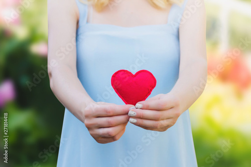 Fototapeta Naklejka Na Ścianę i Meble -  Young woman holding a red heart in her hands against a background of nature. Love, pregnancy, parenthood, motherhood concept, copy space