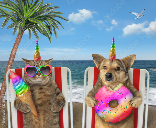 The cat unicorn in sunglasses is eating ice cream cone and dog unicorn is eating a colored donut. They sit under the palm tree on a beach chairs by the sea together. © iridi66