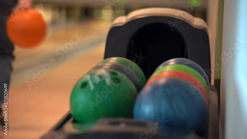 In the frame of the Ball return system with colorful bowling balls, there is a green and blue balls in the background can be seen as a girl in blue jeans throws an orange bowling ball