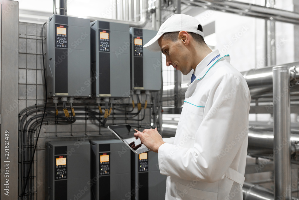 Portrait of man in a white robe and a cap standing in production department of dairy factory with grey tablet Portrait of man in a white robe and a cap standing in production department of dairy factory with grey tablet