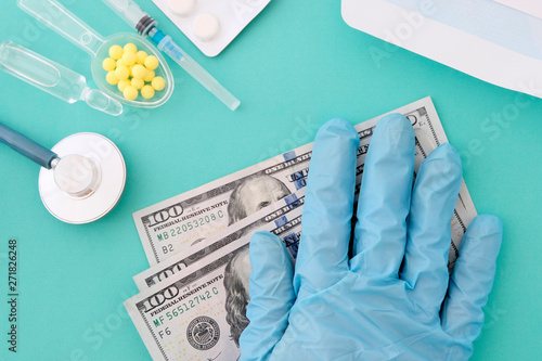 A woman's hand in a blue glove on a hundred-dollar bill. Stethoscope, pills, syringe on the table. The concept of doctor's salary. Blue background.