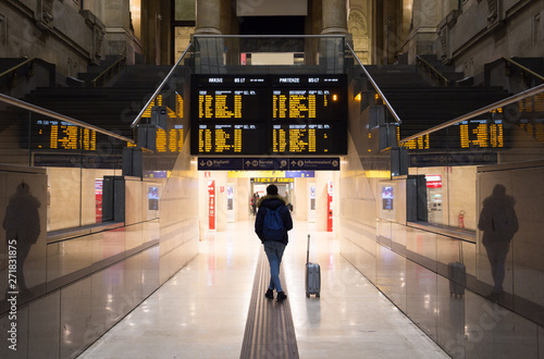 Passenger with a backpack and a suitcase beside her is standing below a timetable board in a giant hall of a railway station in Milan, Italy. Text: 