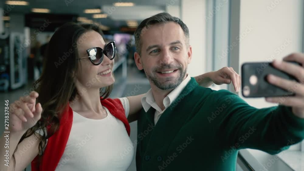 Young beautiful couple in the hall of airport. Brunette smiling woman and man taking selfie photo on smartphone near the window. Cinematic shot on RED camera. Young beautiful couple in the hall of airport. Brunette smiling woman and man taking selfie photo on smartphone near the window. Cinematic shot on RED camera.