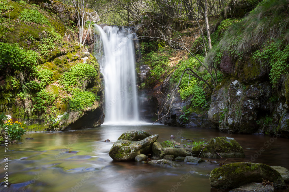 Low perspective view of a scenic mountain waterfall in a vivid green forest lighten by the sun with foreground rocks