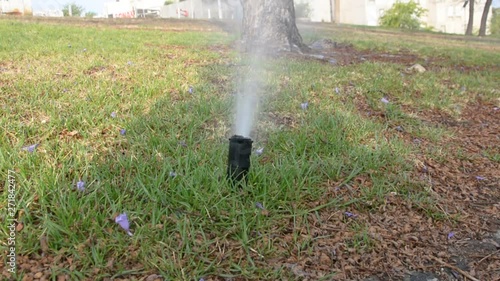 Water sprinkler in public park. Automatic irrigating system watering green grass lawn outdoor. Summer morning. Spray and drops of aqua watering leaves