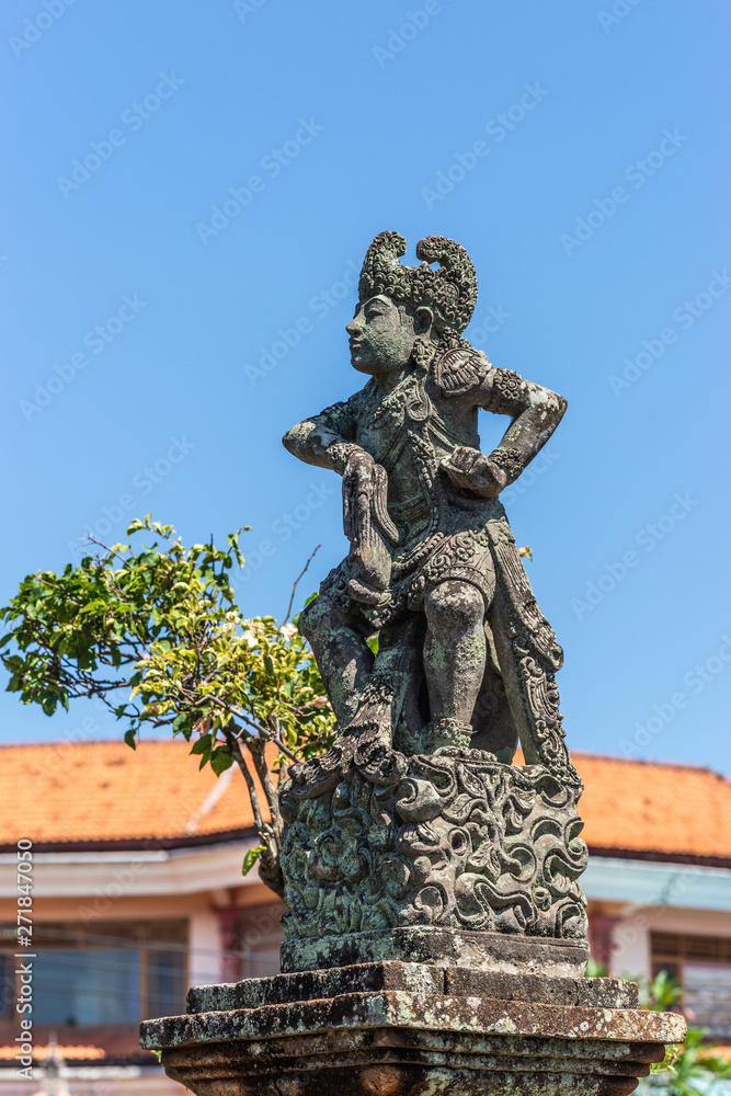 Klungkung, Bali, Indonesia - February 26, 2019: Closeup of Stone statue of defiant King covered in black mold against blue sky. Some green foliage and red roof.