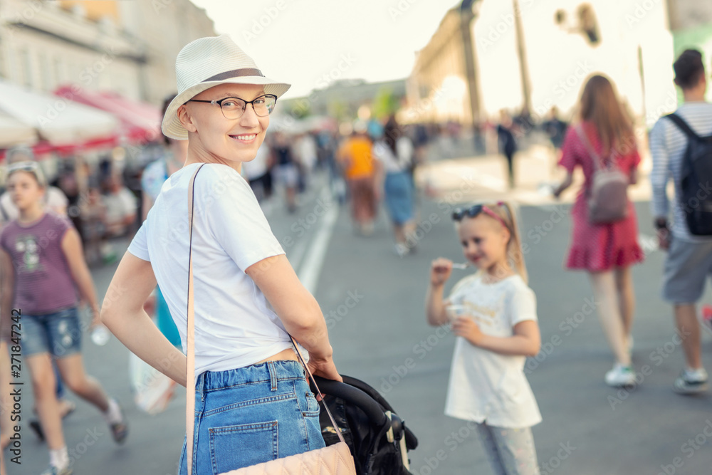 Happy young caucasian bald woman in hat and casual clothes enjoying life  after surviving breast cancer. Portrait of beautiful hairless girl smiling  during walk at city street with after curing disease Stock